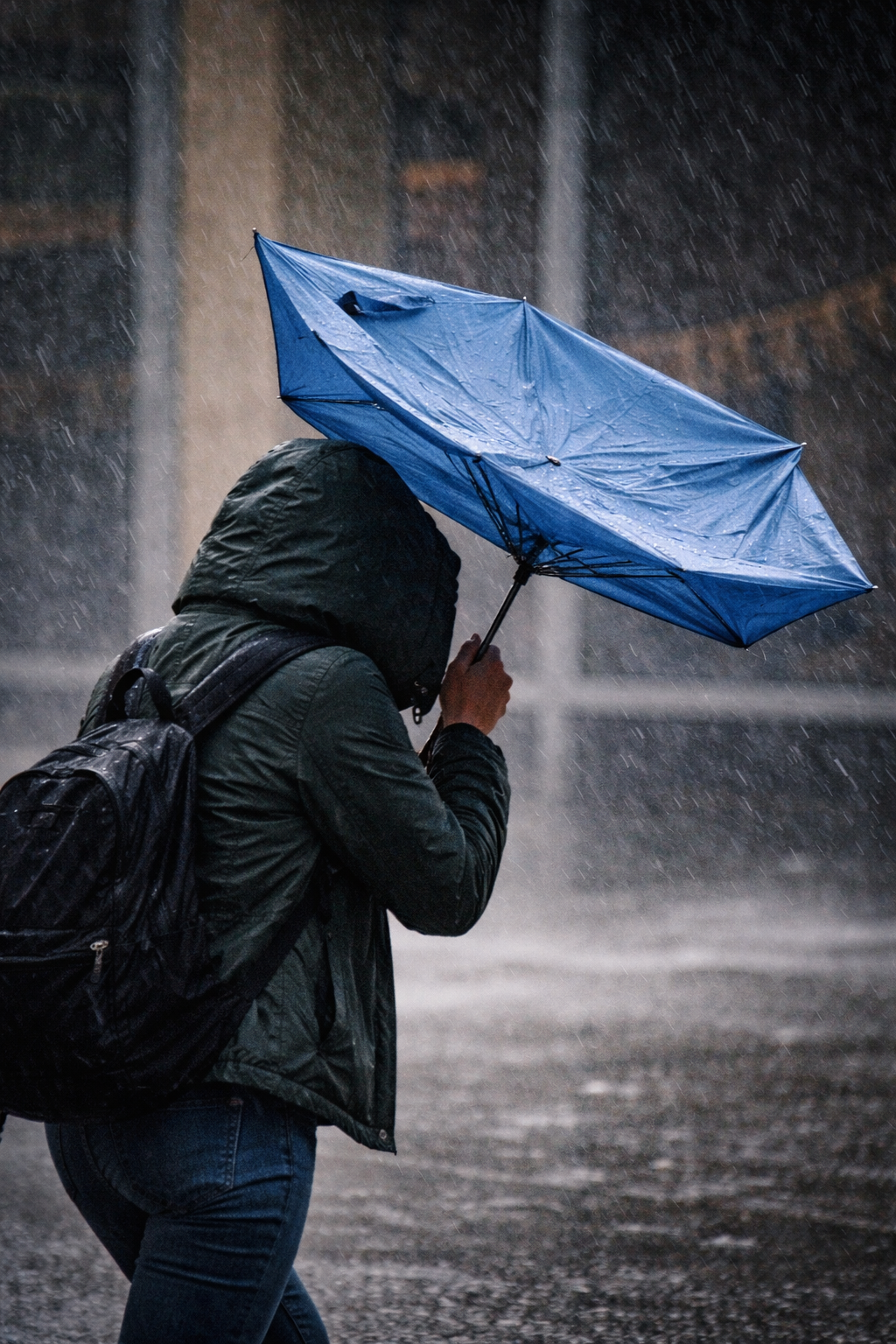 Person in a hooded jacket walking through a heavy storm while an umbrella bends inside out from strong wind.